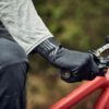 LIN-TERRANOVA-211109-3067-scaled-2.jpg Close-up of a cyclist's gloved hand in a textured black glove gripping a mountain bike handlebar and brake lever, with a grey sleeve and red frame visible.