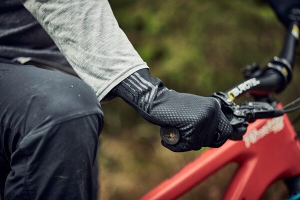 LIN-TERRANOVA-211109-3067-scaled-2.jpg Close-up of a cyclist's gloved hand in a textured black glove gripping a mountain bike handlebar and brake lever, with a grey sleeve and red frame visible.