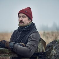 Man in a red knit beanie and brown gloves leans on a weathered wooden gate, wearing a dark jacket and fleece neck gaiter and looking pensive against a foggy field with blurred trees and a lichen‑covered stone post.