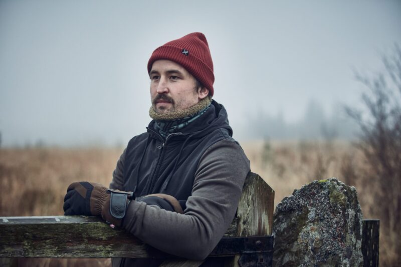 Man in a red knit beanie and brown gloves leans on a weathered wooden gate, wearing a dark jacket and fleece neck gaiter and looking pensive against a foggy field with blurred trees and a lichen‑covered stone post.