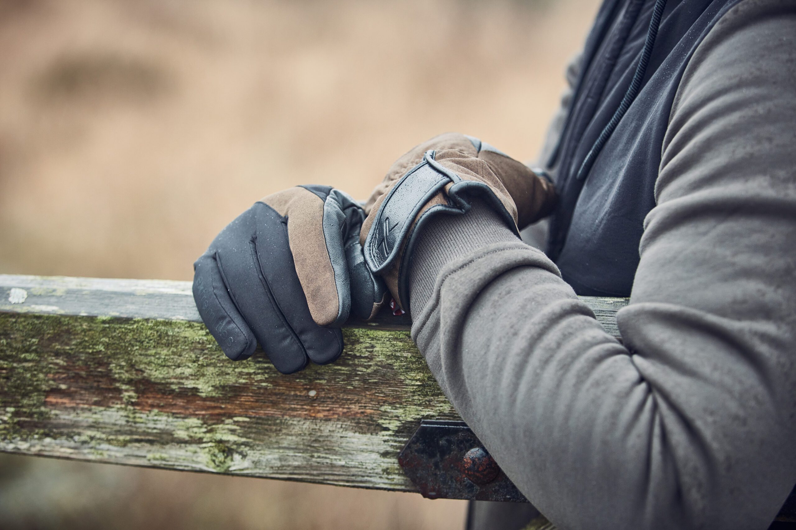 LIN-TERRANOVA-211109-3410-scaled-4.jpg Close-up of a person's brown-and-black insulated gloves resting on a weathered, moss-covered wooden fence rail with a grey hoodie sleeve visible.