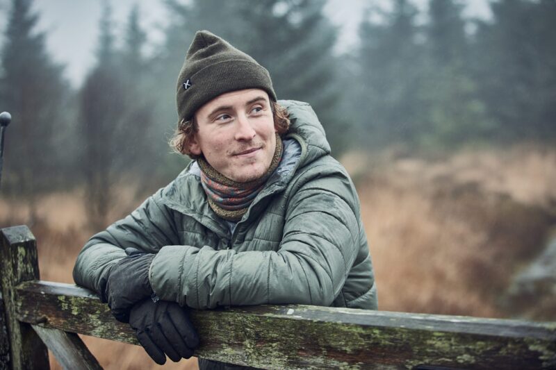Young man in a green beanie and quilted jacket leans on a moss-covered wooden gate in a misty winter landscape, gloved hands crossed on the gate and a patterned neck scarf visible as he looks thoughtfully to the right.