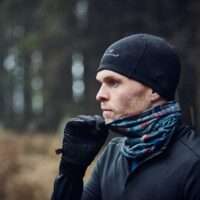 Side-profile of a man wearing a black beanie and gloves, pulling a patterned neck gaiter up over his mouth as rain falls in a dim coniferous woodland.