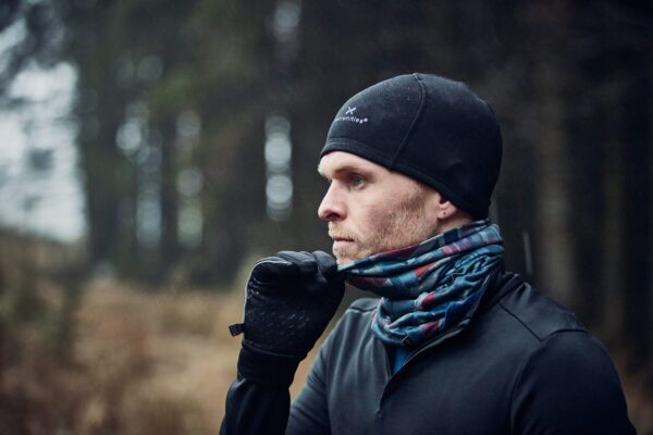 LIN-TERRANOVA-211109-3513-scaled-2.jpg Side-profile of a man wearing a black beanie and gloves, pulling a patterned neck gaiter up over his mouth as rain falls in a dim coniferous woodland.