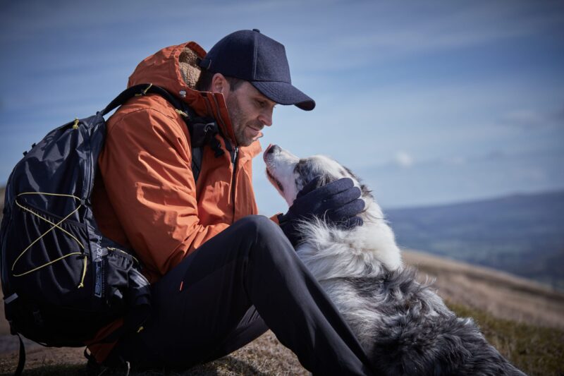 LIN-TERRANOVA-725970-221011-0323-RT-LR-scaled-4.jpg Man in an orange jacket and navy cap sits on a hillside with a backpack, leaning forward to cradle a fluffy white-and-grey dog with gloved hands as the dog lifts its head toward him against a distant blue-sky landscape.