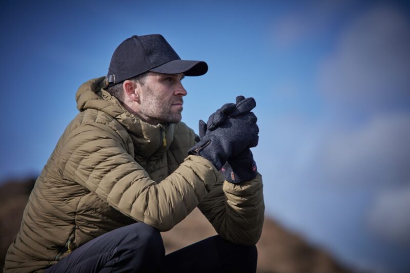 LIN-TERRANOVA-725970-221011-0362-RT-LR-scaled-4.jpg Man in an olive-green puffer jacket and black cap crouches on a rocky hillside, wearing dark gloves and clasping his hands as he gazes into the distance against a clear blue sky.