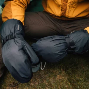Close-up of a person sitting on grass in a mustard-yellow jacket wearing large black insulated mittens with adjustable straps and a visible GORE‑TEX tag, suggesting cold‑weather camping.