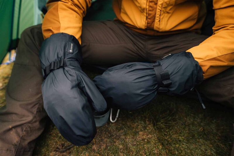 LR-tuff-bags-3.jpg Close-up of a person sitting on grass in a mustard-yellow jacket wearing large black insulated mittens with adjustable straps and a visible GORE‑TEX tag, suggesting cold‑weather camping.