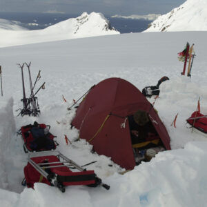 A red camping tent set up in a snowy landscape, surrounded by ski equipment and sleds, with mountains and ocean visible in the background. Ideal for winter mountaineering and outdoor adventures.