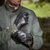 Close-up of a man’s torso and hands as he pulls on black leather gloves over the cuff of an olive-green waxed jacket, his stubbled chin visible and a blurred stone wall and grassy field in the background.