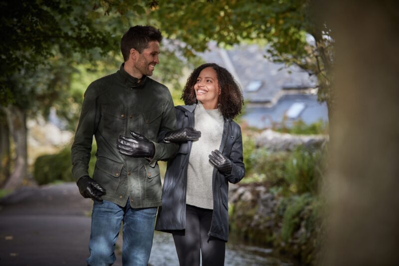 Two people walk arm-in-arm along a tree-lined path beside a narrow stream, smiling and wearing jackets and black leather gloves, with a stone cottage visible in the blurred background.