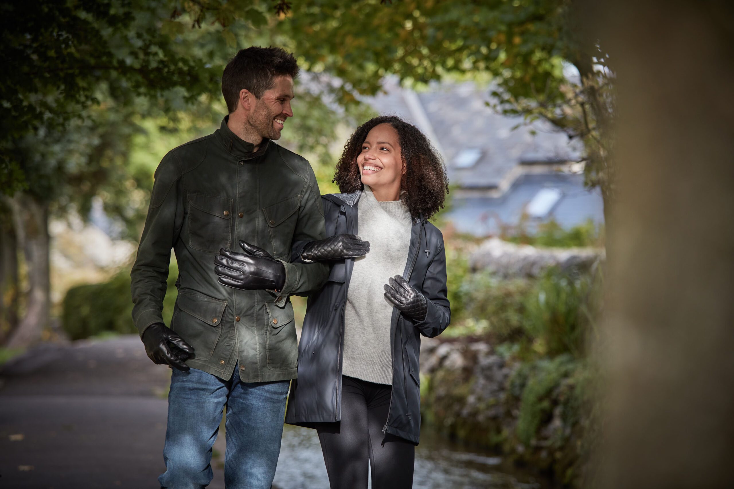 Laax-and-Fernie-Glove-scaled-4.jpg Two people walk arm-in-arm along a tree-lined path beside a narrow stream, smiling and wearing jackets and black leather gloves, with a stone cottage visible in the blurred background.