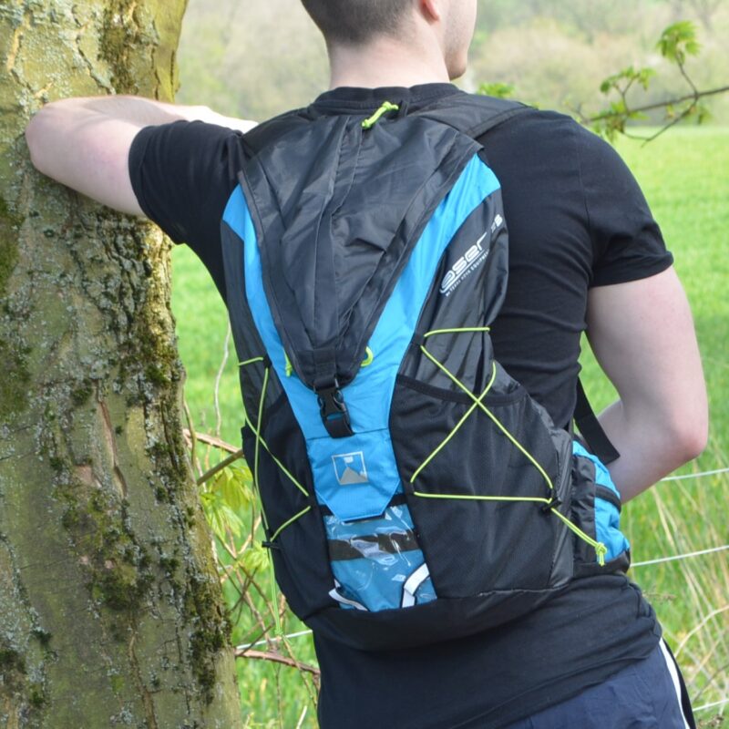 Person seen from behind wearing a black T-shirt and a blue-and-black backpack with neon-green bungee cords, leaning with one arm against a tree in a grassy field.