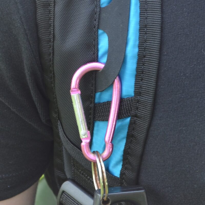Pink metal carabiner clipped to a blue-and-black backpack strap holding a keyring and keys in a close-up of a person's shoulder.