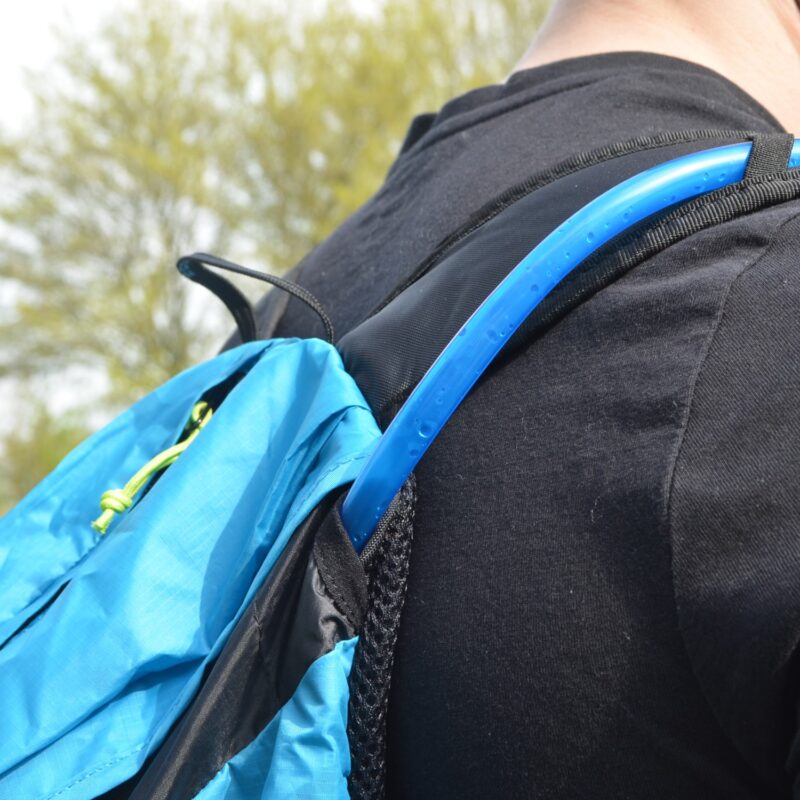 Close-up of a person's shoulder in a black t-shirt wearing a bright blue backpack with a blue hydration tube draped over the strap, water droplets on the tube and a neon green zipper pull visible, with blurred trees in the background.