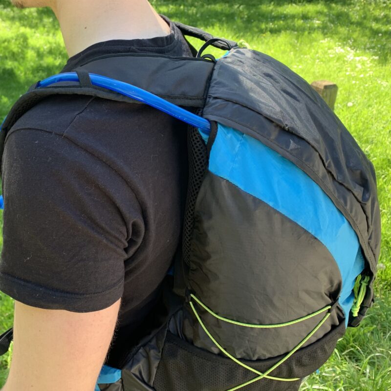 Side view of a person in a black t-shirt wearing a black-and-blue hiking backpack with a blue hydration tube over the shoulder and neon-green bungee cords, against a grassy background.