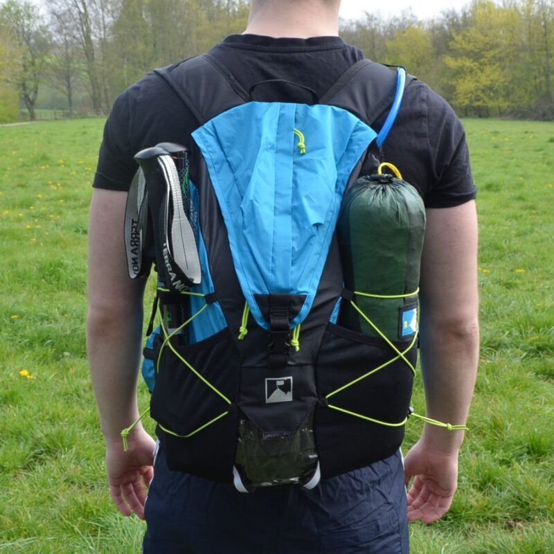Rear view of a person wearing a blue-and-black hiking backpack with neon-yellow bungee cords, a rolled green sleeping bag strapped to the side, trekking poles tucked into the left pocket and a blue hydration tube over the shoulder, standing in a grassy field.