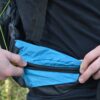 Laser-25-side-pocket-1500x1500-1-1.jpg Two hands opening a small bright blue zippered pocket on a black backpack hip belt worn over a black shirt, showing ripstop nylon fabric and a black zipper.