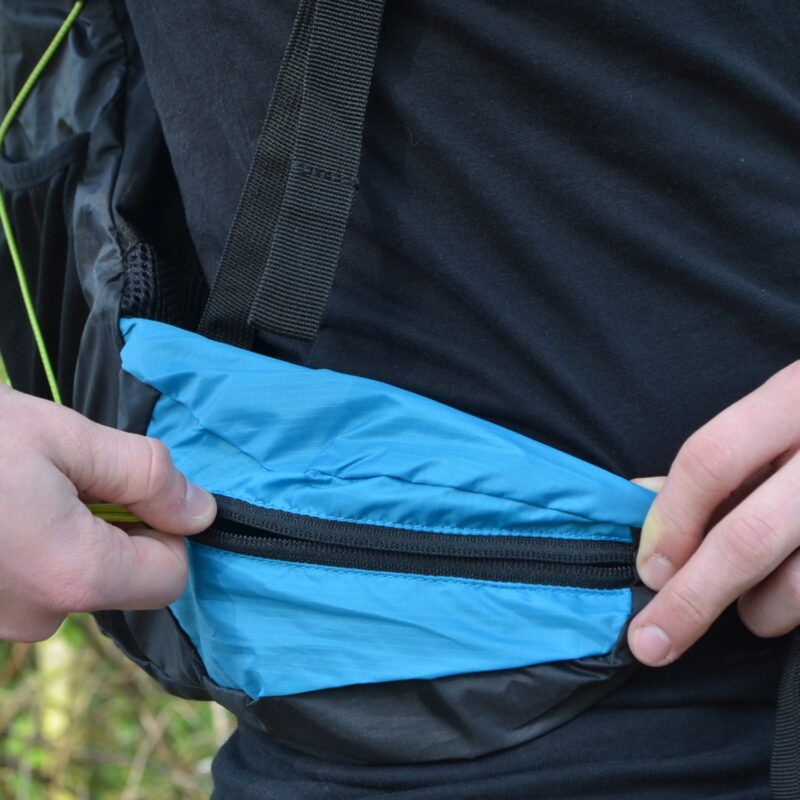 Two hands opening a small bright blue zippered pocket on a black backpack hip belt worn over a black shirt, showing ripstop nylon fabric and a black zipper.