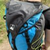 Laser-35-top-view-1500px-1.jpg Back view of a person wearing a bright blue and black hiking backpack with a folded top flap, visible straps and buckles and a neon-yellow cord, photographed outdoors on a wooded trail.