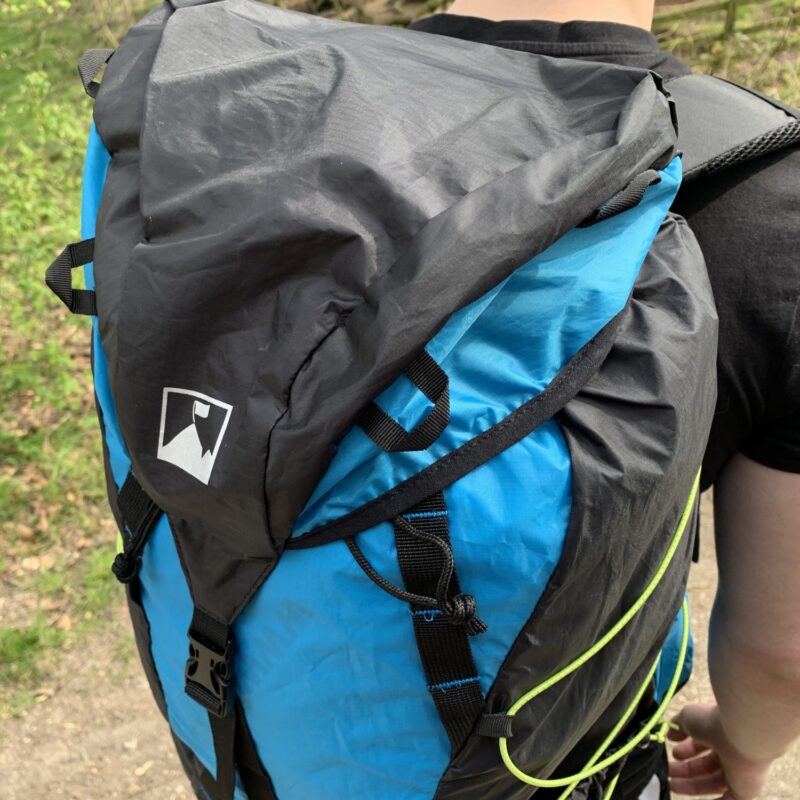 Laser-35-top-view-1500px-1.jpg Back view of a person wearing a bright blue and black hiking backpack with a folded top flap, visible straps and buckles and a neon-yellow cord, photographed outdoors on a wooded trail.