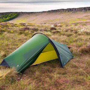 A small green two-person tent with a yellow inner lining staked on windswept heather and grass moorland dotted with white cotton-grass and yellow gorse, set against a low rocky escarpment and an overcast sky.