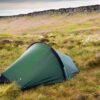A small green dome tent staked with yellow guy lines on windswept grassy moorland dotted with white wildflowers, with a rugged rocky escarpment and cloudy sky in the background.