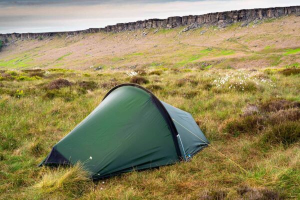 A small green dome tent staked with yellow guy lines on windswept grassy moorland dotted with white wildflowers, with a rugged rocky escarpment and cloudy sky in the background.