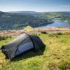 Small dark-coloured backpacking tent pitched on a grassy hilltop overlooking a tree-lined reservoir and dam with rolling green hills and a clear blue sky.