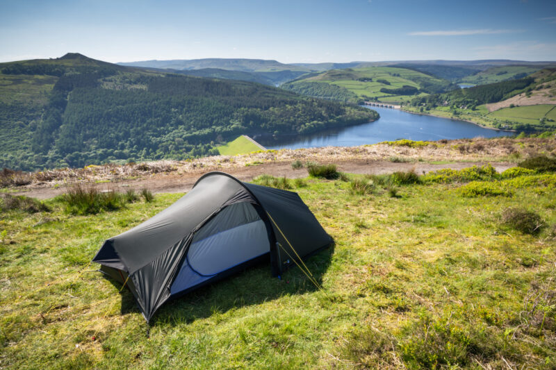 Small dark-coloured backpacking tent pitched on a grassy hilltop overlooking a tree-lined reservoir and dam with rolling green hills and a clear blue sky.