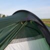 Laser-Compact-All-Season-2-Lifestyle-Door-Hood-1500x1500px-1.jpg Close-up of a green camping tent's partially open entrance showing a dark rainfly with yellow toggles and a light grey inner panel, set against a grassy field and pale blue sky.