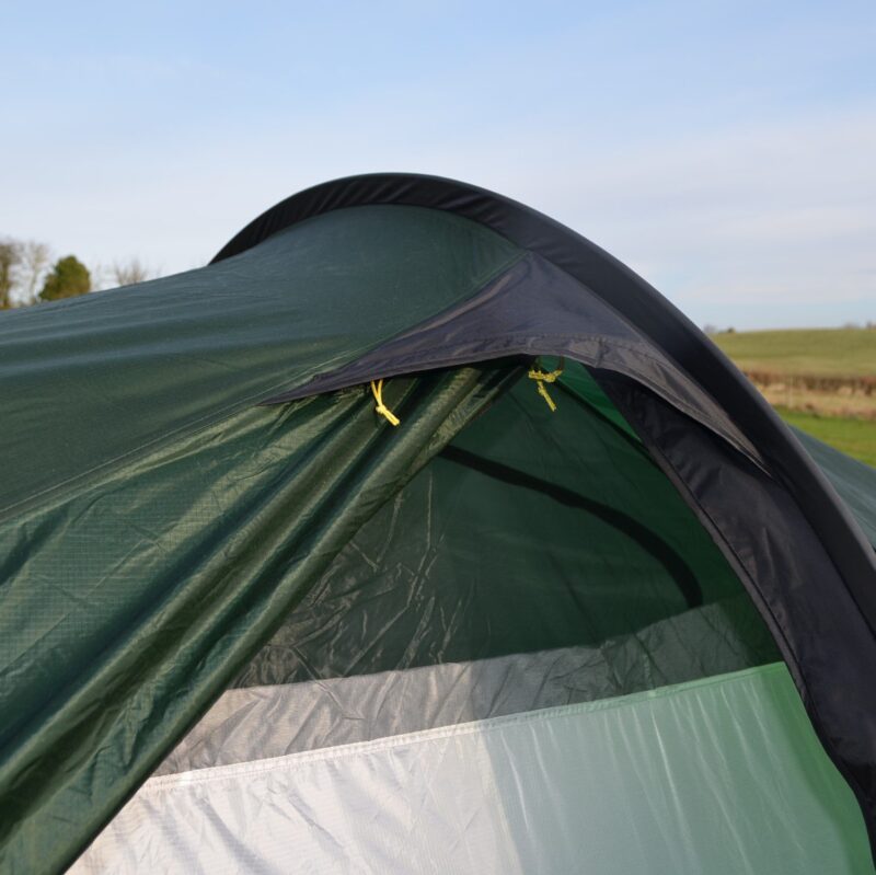Laser-Compact-All-Season-2-Lifestyle-Door-Hood-1500x1500px-1.jpg Close-up of a green camping tent's partially open entrance showing a dark rainfly with yellow toggles and a light grey inner panel, set against a grassy field and pale blue sky.