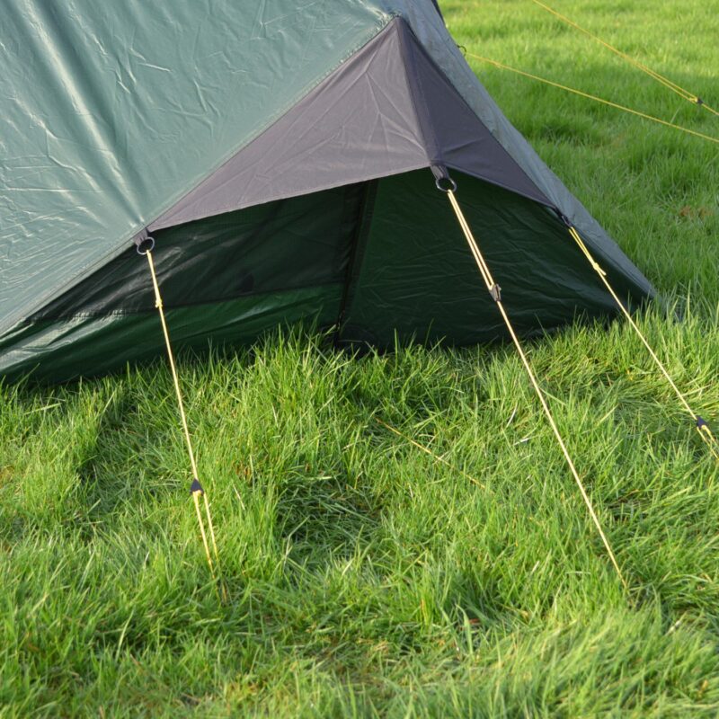 Laser-Compact-All-Season-2-Lifestyle-Extra-End-Guylines1500x1500px-1.jpg Close-up of a green tent's lower corner with a dark grey triangular vent flap held open by yellow guy lines and metal rings, staked into lush green grass.