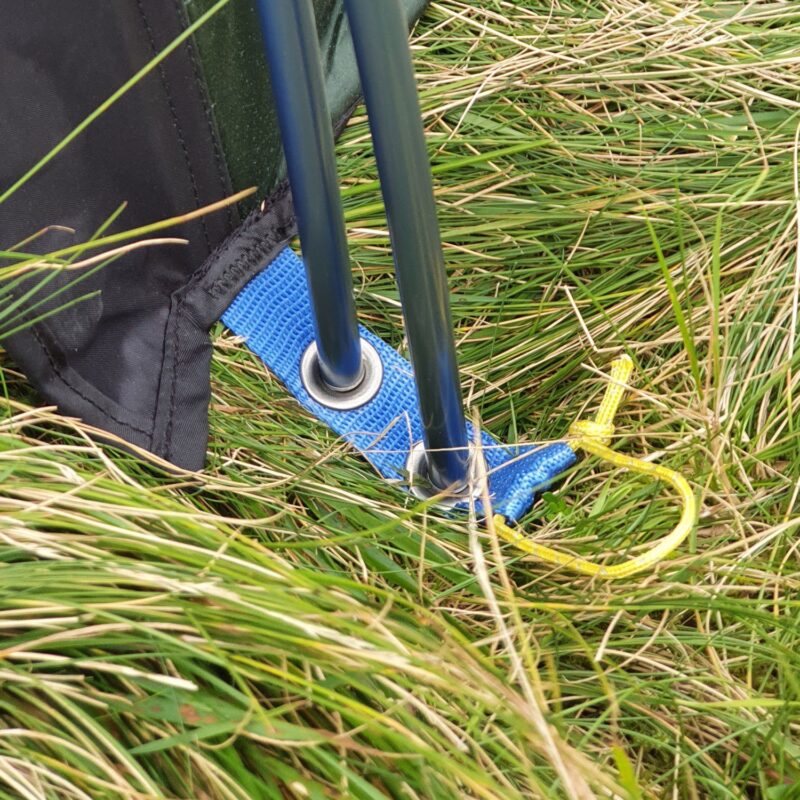 Laser-Compact-All-Season-option-to-double-pole-1500x1500-1-1.jpg Close-up of two dark metal tent poles passing through a blue webbing loop with metal eyelets and a tied yellow cord, set among long green grass beside black fabric
