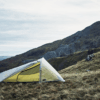 Laser-Pulse-Ultra-1-1.png Lightweight yellow one-person tent pitched on a windswept grassy hillside with rocky mountains and an overcast sky in the background