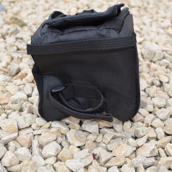 Laser-Velo-Frame-Back-Attachment-1.jpg Small black padded pouch with a zippered top and Velcro strap, lying on pale loose gravel with a short looped side handle visible.