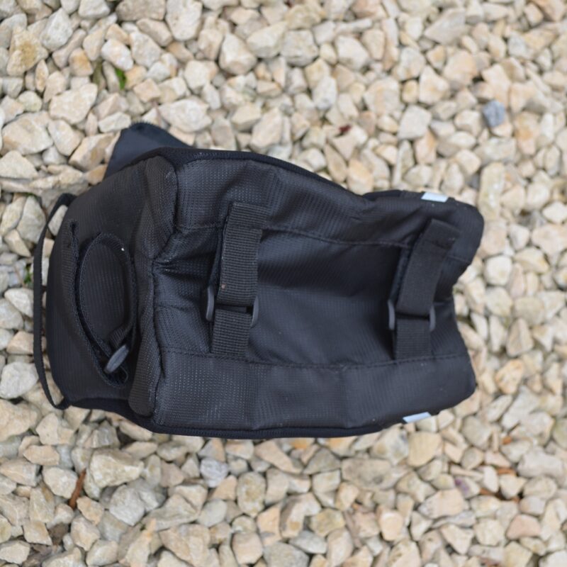 Black nylon pouch with two vertical strap loops and a rounded side pocket, seen from above lying on pale gravel.