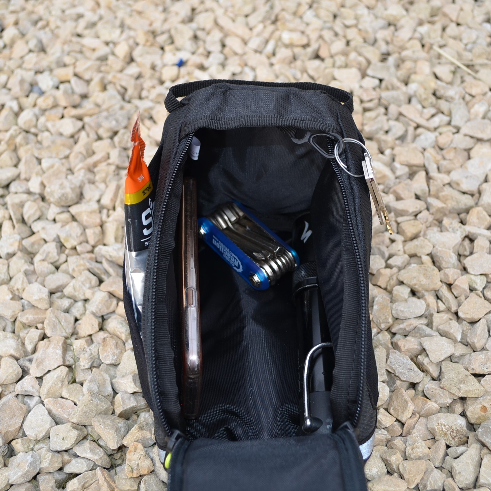 Laser-Velo-Frame-Showing-Space-Inside-1.jpg Open black zippered pouch on gravel containing a smartphone, a blue multitool, an orange energy gel sachet tucked into the side, a key on a metal ring clipped to the zipper and a small black pump with a metal clip.