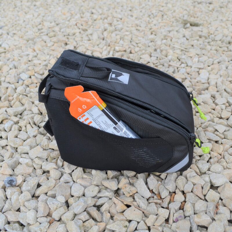 Small black zippered pouch with neon-green zipper pulls and a white logo, a bright orange packet peeking from its mesh side pocket, resting on pale gravel.