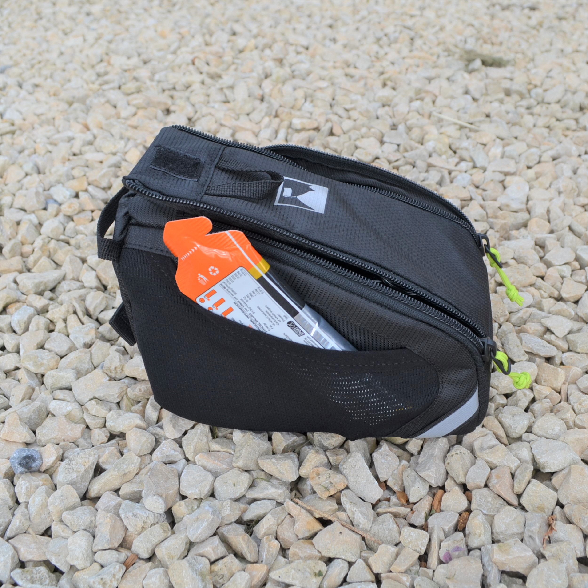 Laser-Velo-Frame-Side-View-Pocket-1.jpg Small black zippered pouch with neon-green zipper pulls and a white logo, a bright orange packet peeking from its mesh side pocket, resting on pale gravel.