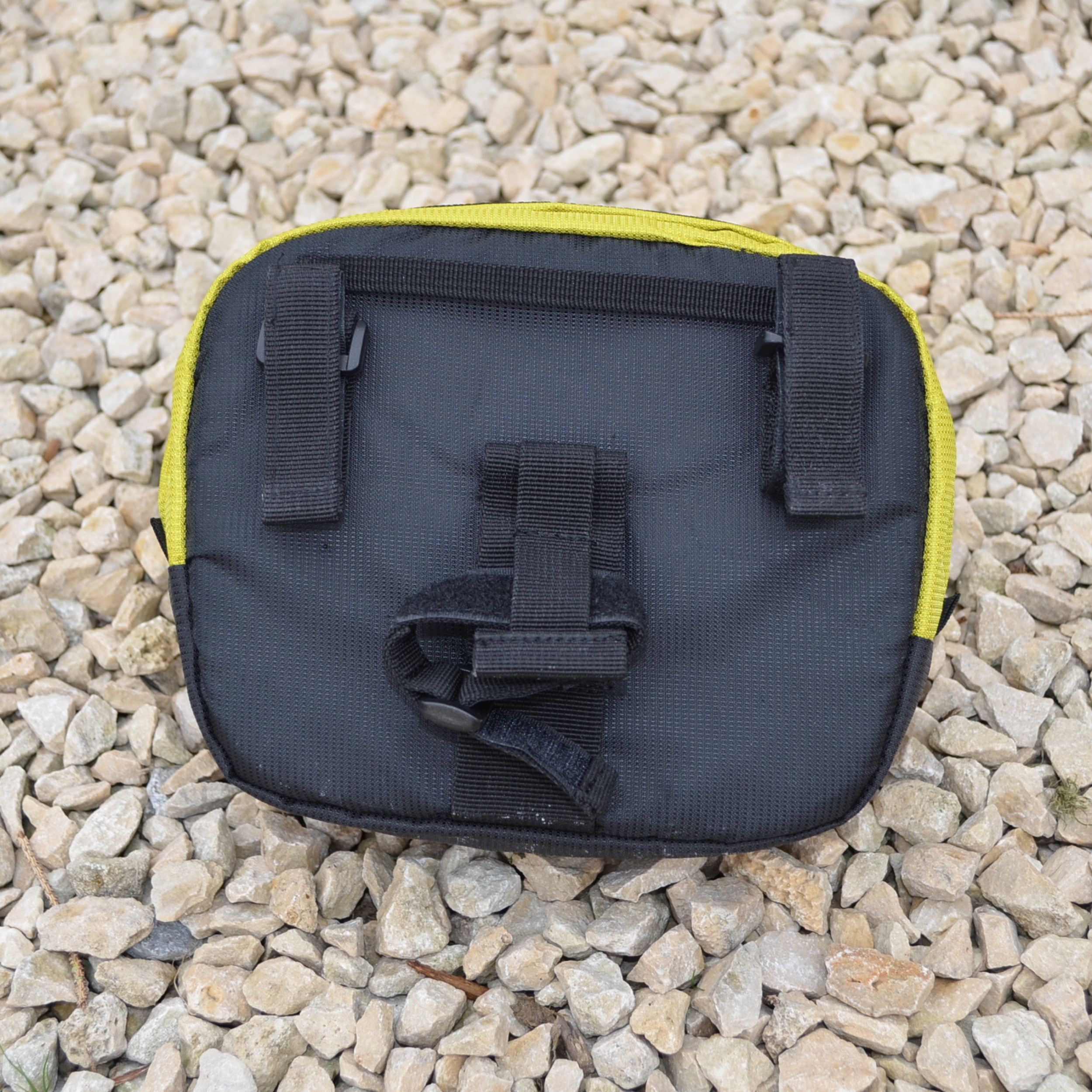 Laser-Velo-Handlebar-Attachment-Points-1.jpg Small rectangular black fabric pouch with yellow trim, two vertical belt loops and a central velcro strap visible on its back, lying on a bed of pale gravel.