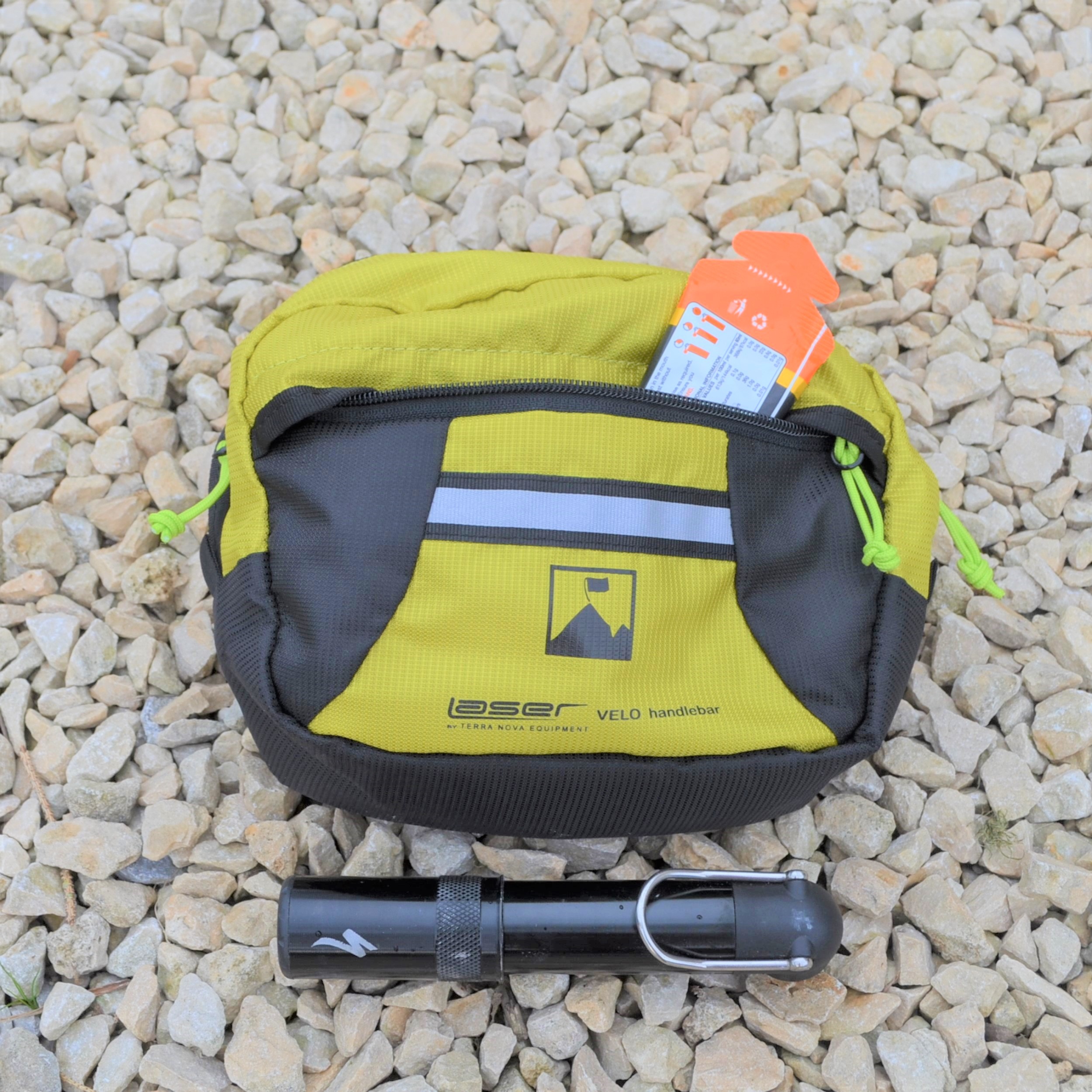 Laser-Velo-Handlebar-Front-Pocket-Detail-1.jpg Yellow-green and grey Velo handlebar bag with an orange package peeking from its zippered pocket, resting on pale gravel with a small black bicycle pump placed in front.