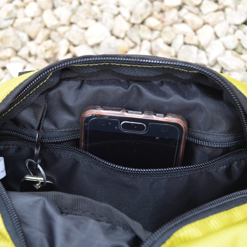 Laser-Velo-Handlebar-Inside-Pocket-Close-Up-1.jpg A black smartphone in a clear protective case sticks out of a zippered inner pocket of a yellow fabric pouch, a key on a ring peeks from the pocket and pale gravel is visible in the background.