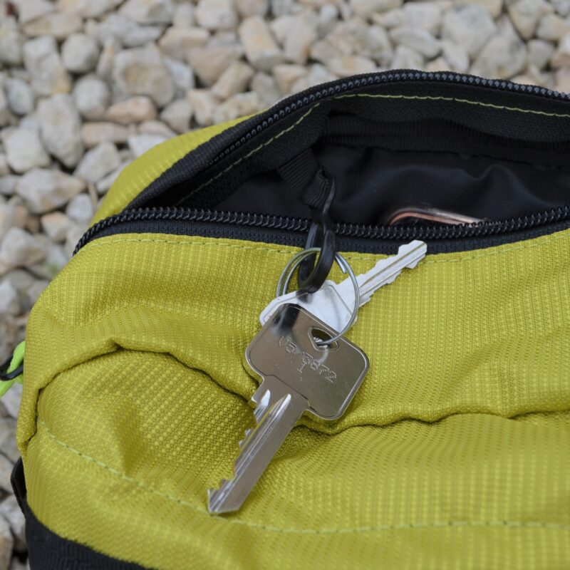 Laser-Velo-Handlebar-Key-Attachment-1.jpg Silver house key on a small keyring lies on a partially open lime-green zippered pouch, with a smaller key and black zipper visible against pale gravel.