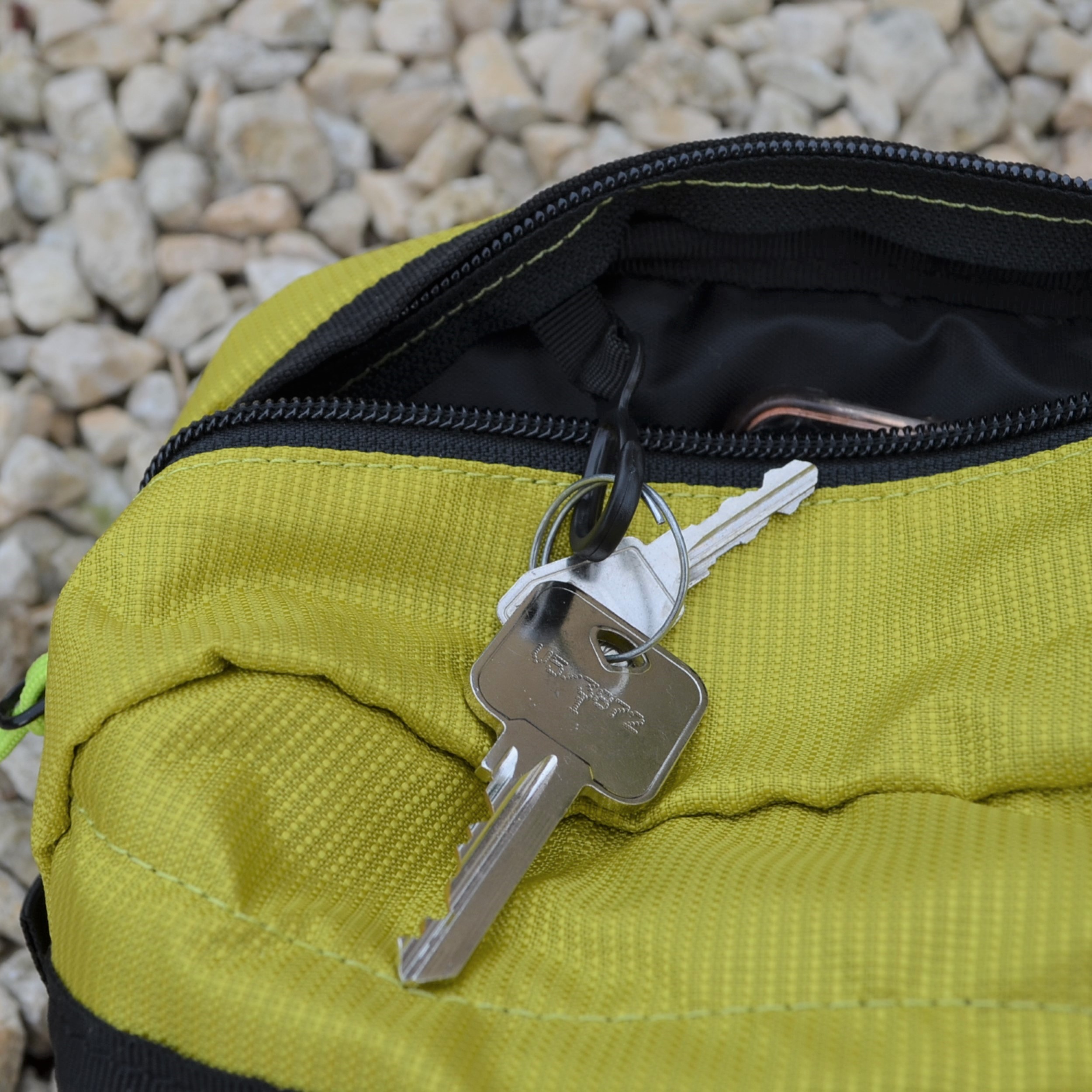 Laser-Velo-Handlebar-Key-Attachment-1.jpg Silver house key on a small keyring lies on a partially open lime-green zippered pouch, with a smaller key and black zipper visible against pale gravel.