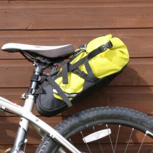 Bicycle saddle with a bright yellow saddlebag secured by black straps beneath the seat above the rear tyre, set against a wooden panel wall.