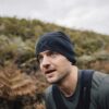Close-up of a man with light stubble wearing a black beanie with a subtle logo and an olive-green top, leaning forward with backpack straps visible and gazing upward against a blurred hillside of ferns and an overcast sky.