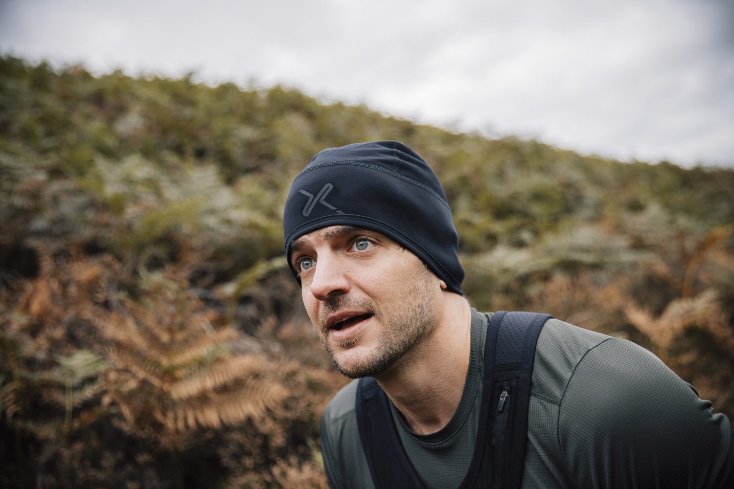 Maze-Runner-Beanie-3.jpg Close-up of a man with light stubble wearing a black beanie with a subtle logo and an olive-green top, leaning forward with backpack straps visible and gazing upward against a blurred hillside of ferns and an overcast sky.