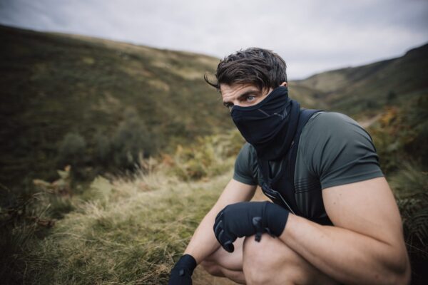 Maze-Runner-Face-Mask-2-3.jpg A man in a short-sleeved green top, black neck gaiter and gloves crouches on a grassy hillside, gazing to the side with rolling hills and a cloudy sky behind him.