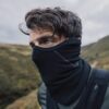 Maze-Runner-Face-Mask-9-3.jpg Close-up portrait of a man with tousled brown hair and blue eyes wearing a black neck gaiter and outdoor gear on a windswept grassy hillside under a cloudy sky